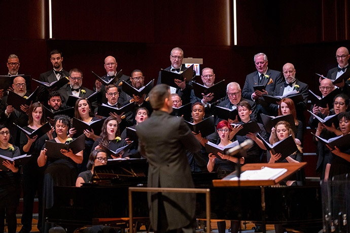WCPA members sing on stage, they are dressed in black clothing and hold song  books. The conductor is in the forefront of the photo.